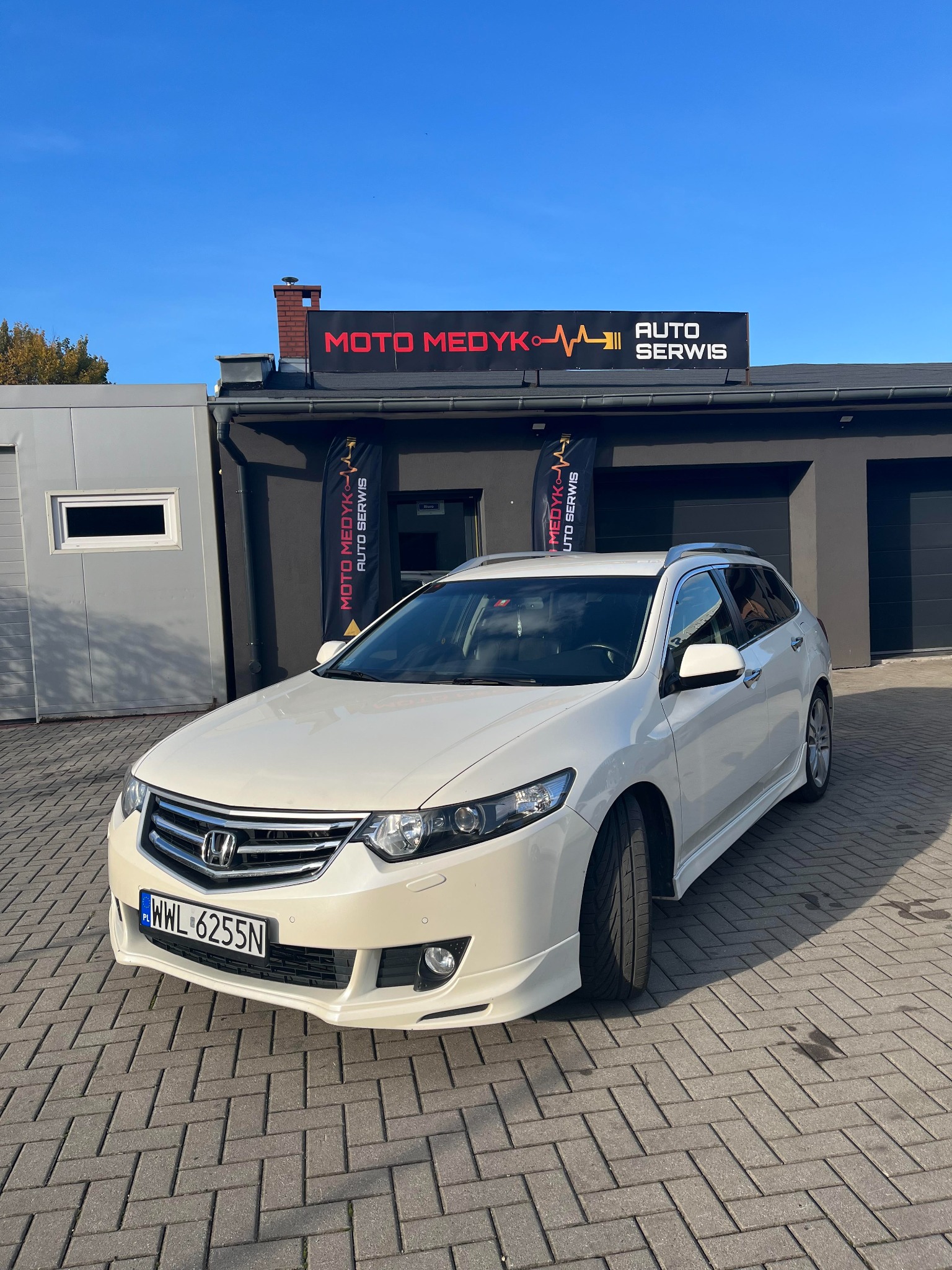 Białe kombi Honda Accord z pakietem stylistycznym stoi przed budynkiem warsztatu Moto Medyk Auto Serwis z czarnym szyldem i flagami reklamowymi.
