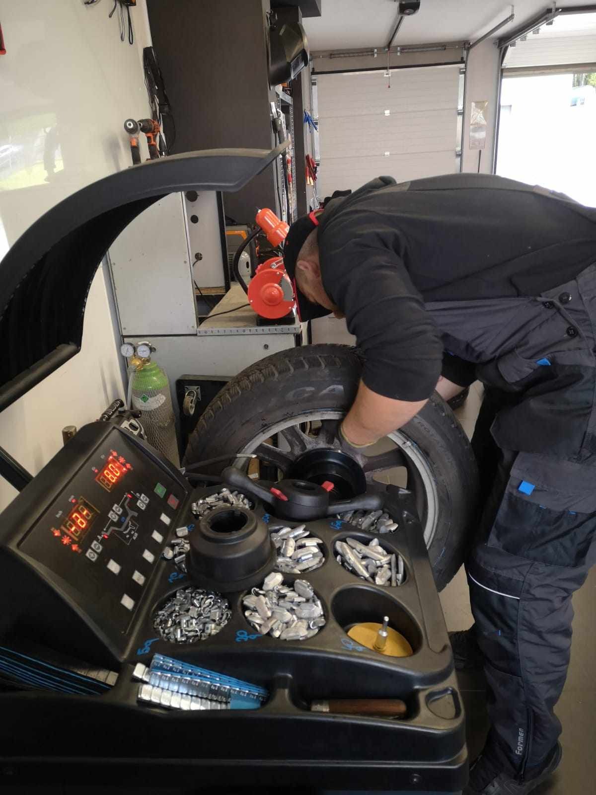 Mężczyzna w warsztacie samochodowym wyważa koło na maszynie do wyważania, widoczne ciężarki i narzędzia.