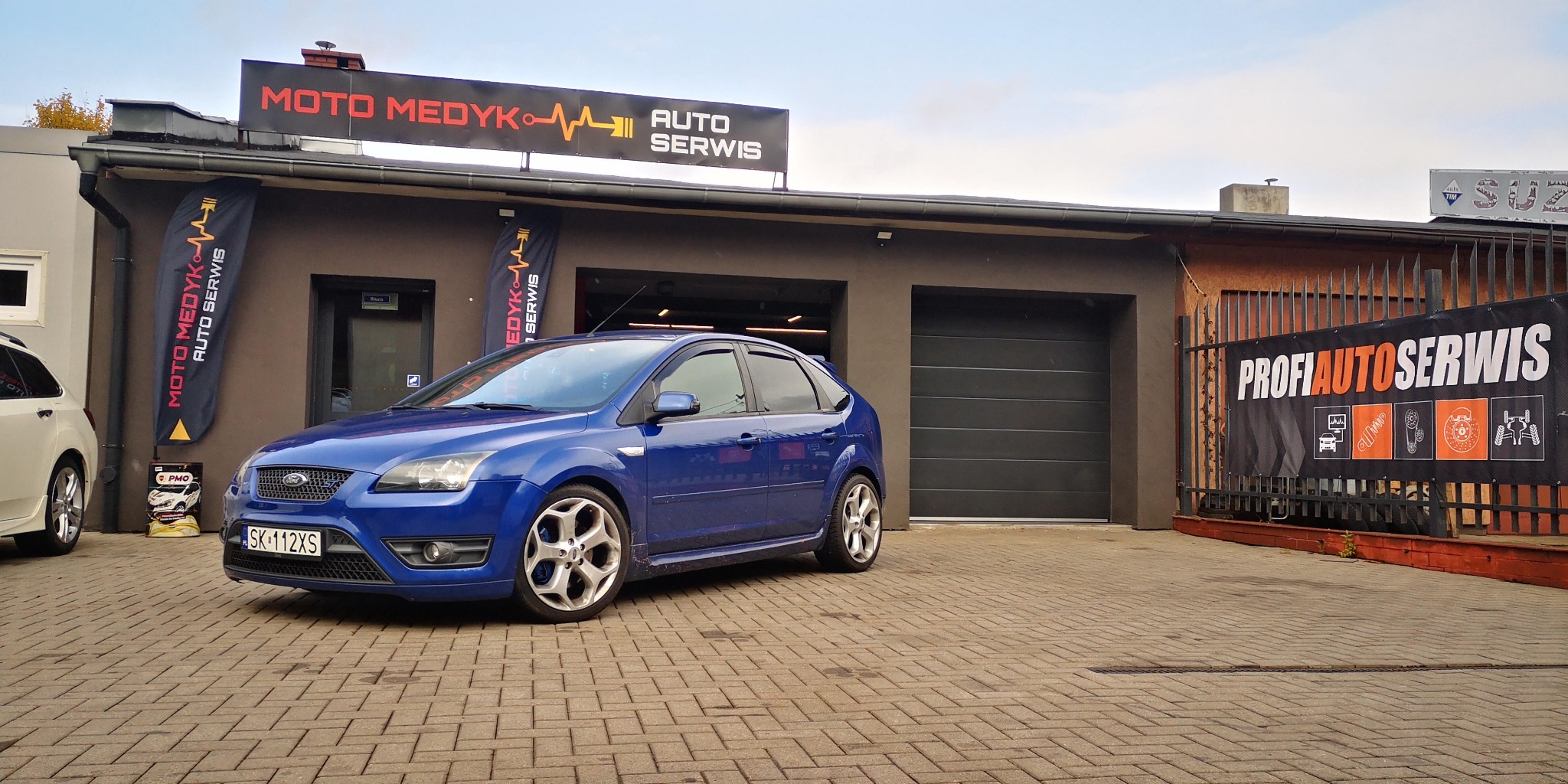 Niebieski Ford Focus zaparkowany przed warsztatem samochodowym Moto Medyk Auto Serwis, widoczny szyld i flaga reklamowa, w tle brama garażowa oraz baner ProfiAutoSerwis.
