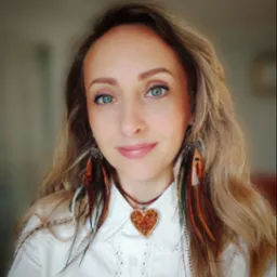 Portret kobiety z blond włosami i niebieskimi oczami, noszącej kolczyki z piór i naszyjnik w kształcie serca z bursztynu, w białej koszuli z koronkowym wykończeniem.