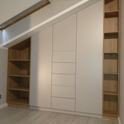 Ww Meble Comfort - Biała szafa wnękowa z drewnianymi półkami, dopasowana do skosu dachu, z widocznymi szufladami, oświetleniem i drewnianą podłogą.