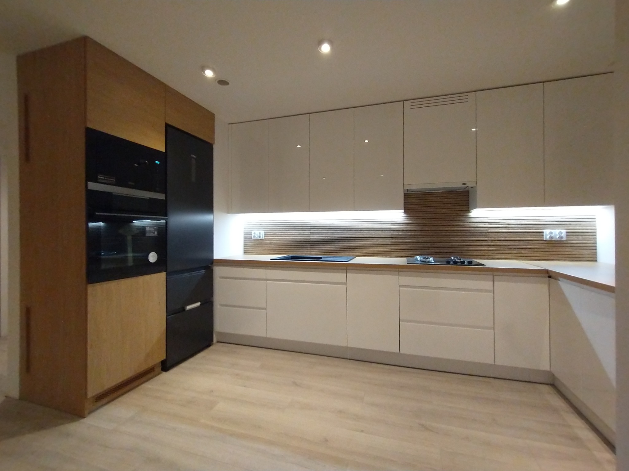 Nowoczesna, jasna kuchnia z drewnianymi akcentami: zabudowa z frontami na wysoki połysk, czarny sprzęt AGD w zabudowie, drewniana ściana nad blatem, oświetlenie LED pod szafkami wiszącymi.