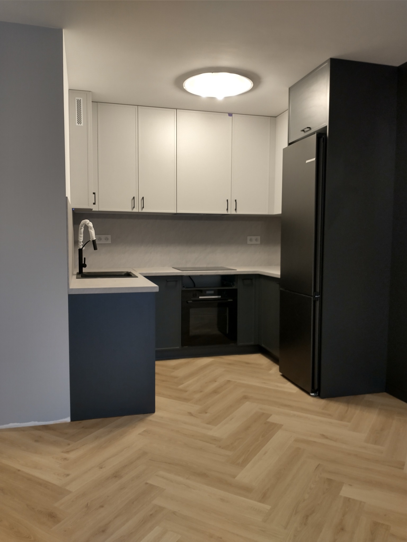 Nowoczesna kuchnia z białymi szafkami górnymi i granatowymi dolnymi, czarny zlew i bateria, duża czarna lodówka, parkiet w jodełkę.