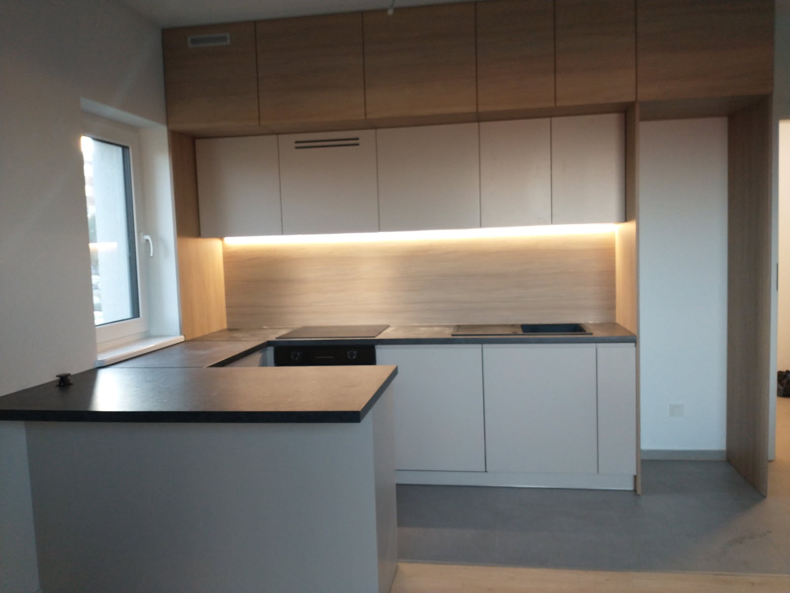 Nowoczesna kuchnia z wyspą, jasne szafki dolne, drewnopodobne szafki górne, oświetlenie LED pod szafkami, czarny blat, widok na okno.