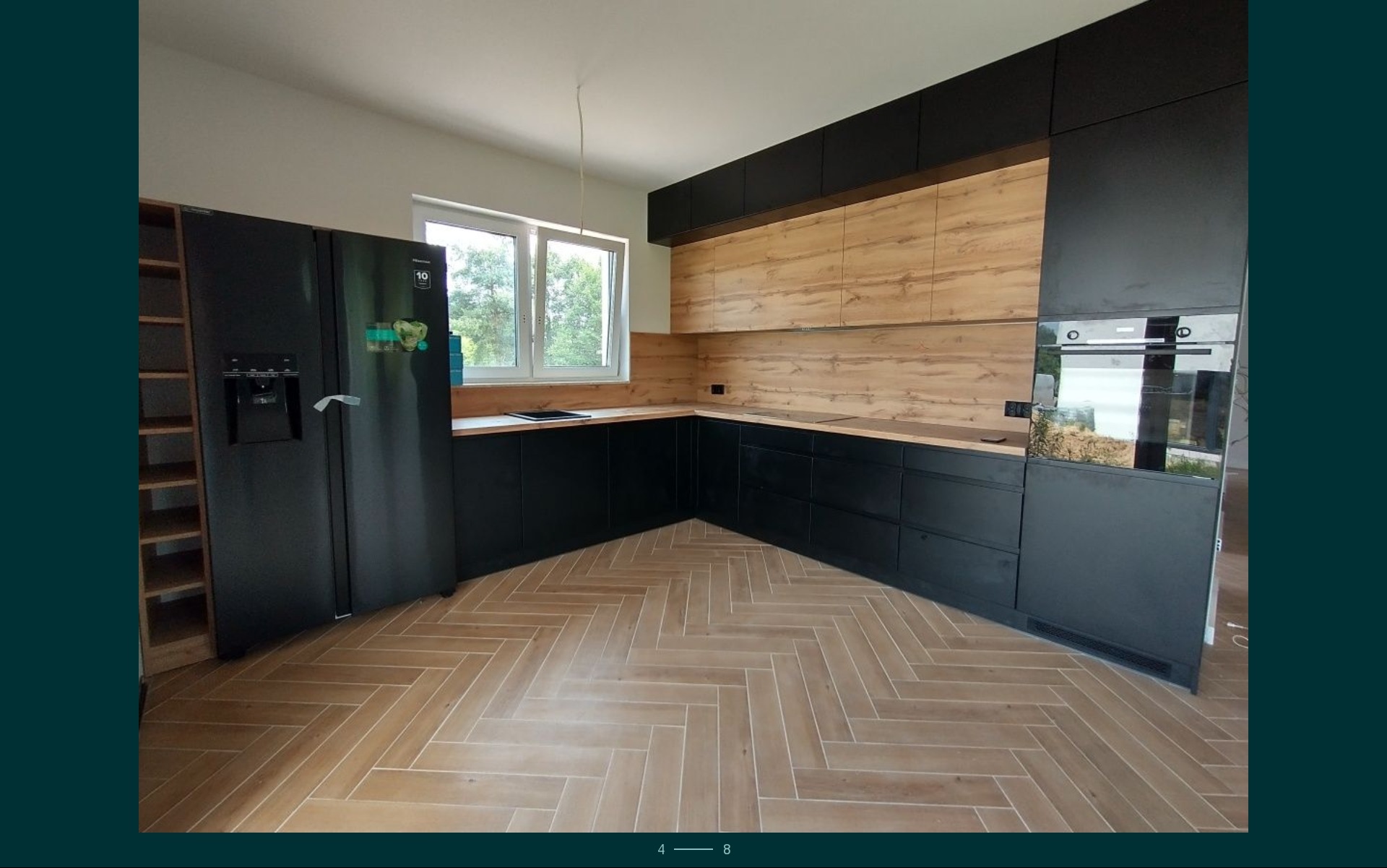 Nowoczesna kuchnia w czerni i drewnie, z zabudową od podłogi do sufitu, drewnianym blatem i podłogą w jodełkę, duża lodówka na lewo od okna.