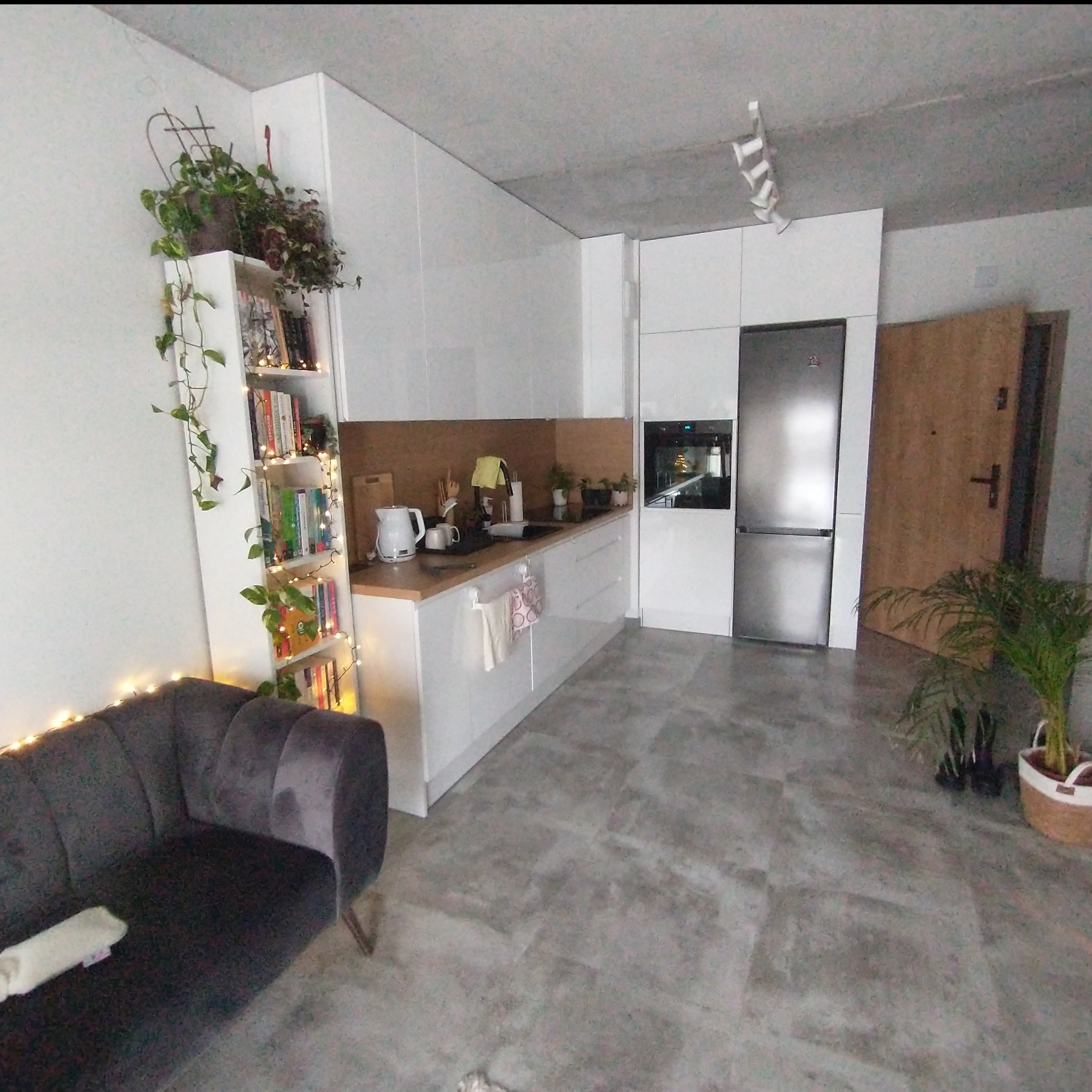 Nowoczesna kuchnia z białymi frontami, drewnianym blatem i backsplash'em, oświetlona półka z książkami i roślinami pnącymi, szary welurowy narożnik z lampkami dekoracyjnymi, betonowa podłoga.