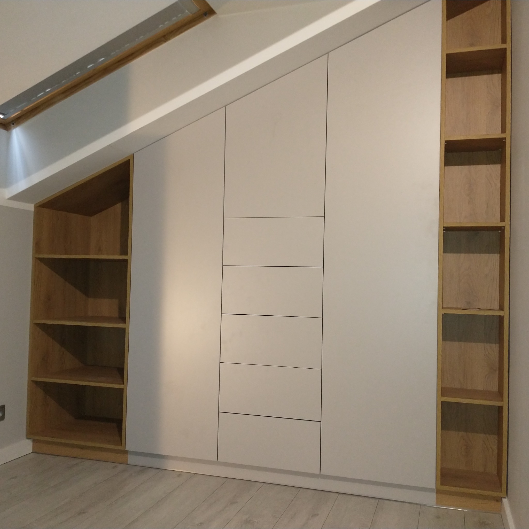 Biała szafa wnękowa z drewnianymi półkami, dopasowana do skosu dachu, z widocznymi szufladami, oświetleniem i drewnianą podłogą.