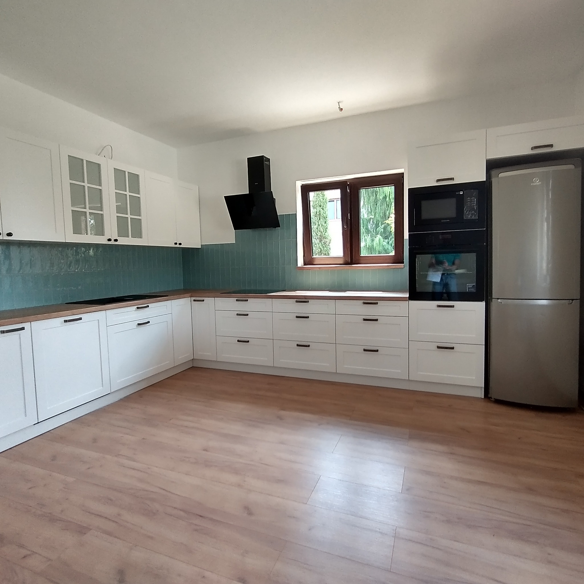 Nowoczesna kuchnia z białymi frontami szafek, drewnianym blatem i turkusowymi płytkami na ścianie, z widocznym oknem i sprzętem AGD.