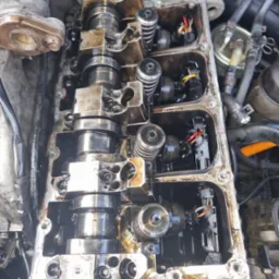 Otwarty silnik samochodowy z widocznymi wałkami rozrządu, sprężynami zaworowymi i przewodami elektrycznymi, pokryty olejem, sugerujący prace naprawcze lub diagnostyczne.