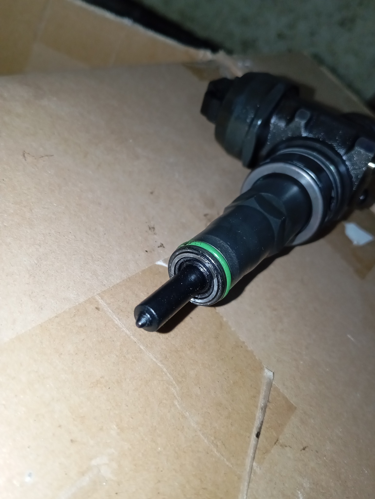 Czarny wtryskiwacz paliwa z zielonym pierścieniem uszczelniającym, leżący na tekturowym tle w warsztacie w Żarach.