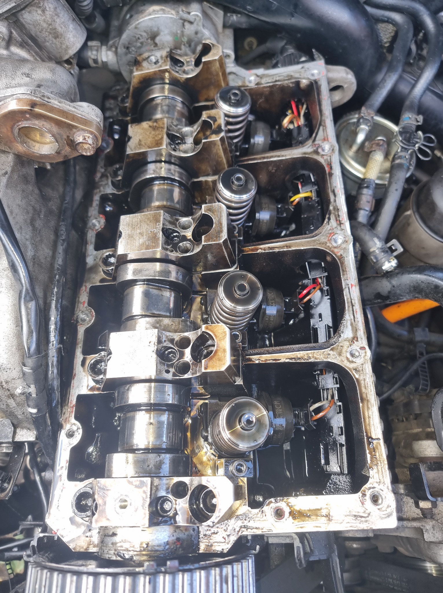 Otwarty silnik samochodowy z widocznymi wałkami rozrządu, sprężynami zaworowymi i przewodami elektrycznymi, pokryty olejem, sugerujący prace naprawcze lub diagnostyczne.