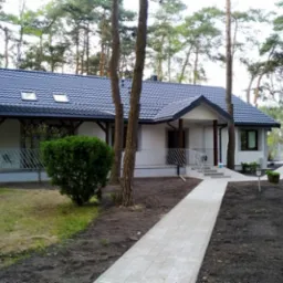 Nowoczesny dom jednorodzinny z szarą dachówką i białą elewacją, otoczony lasem sosnowym, z brukowanym chodnikiem prowadzącym do wejścia i świeżo zagospodarowanym terenem wokół