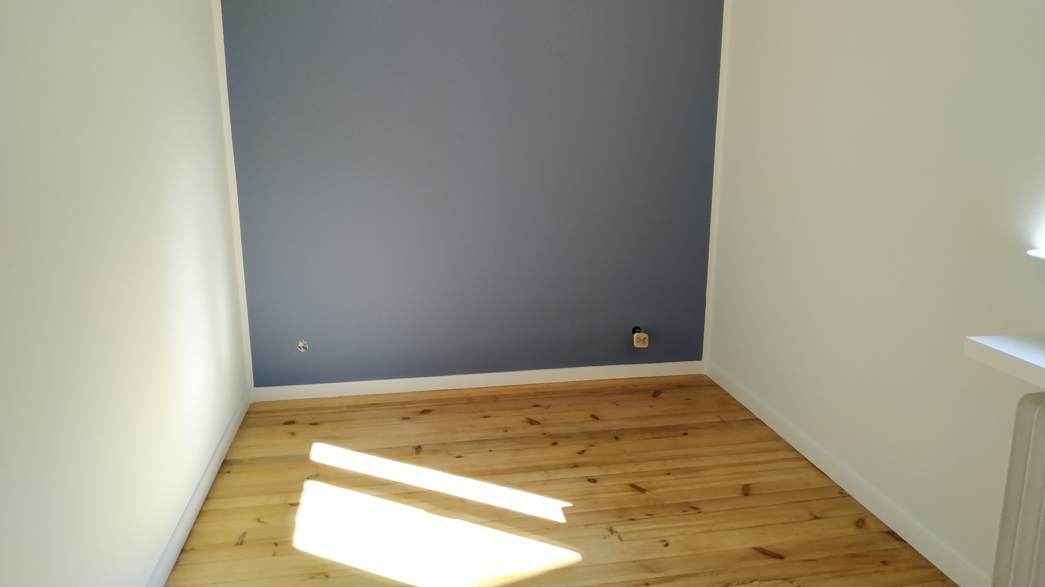 Pomalowane na szaro wnętrze pokoju z drewnianą podłogą, widoczne gniazdka elektryczne w ścianie, białe listwy przypodłogowe i fragment białego grzejnika.