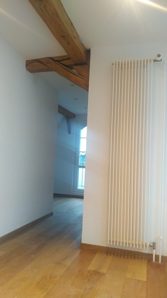 Wnętrze z drewnianą podłogą, białymi ścianami, grzejnikiem panelowym i fragmentem drewnianej belki stropowej, widok na okno w oddali.