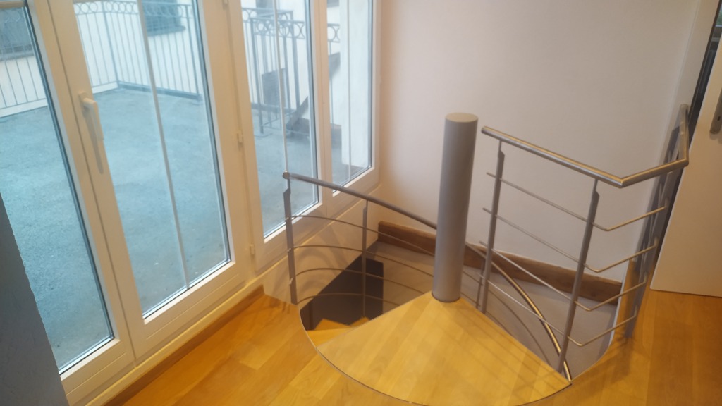 Wnętrze z drewnianymi schodami spiralnymi z metalową balustradą, widok z góry na jasne podłogi i duże okno balkonowe.