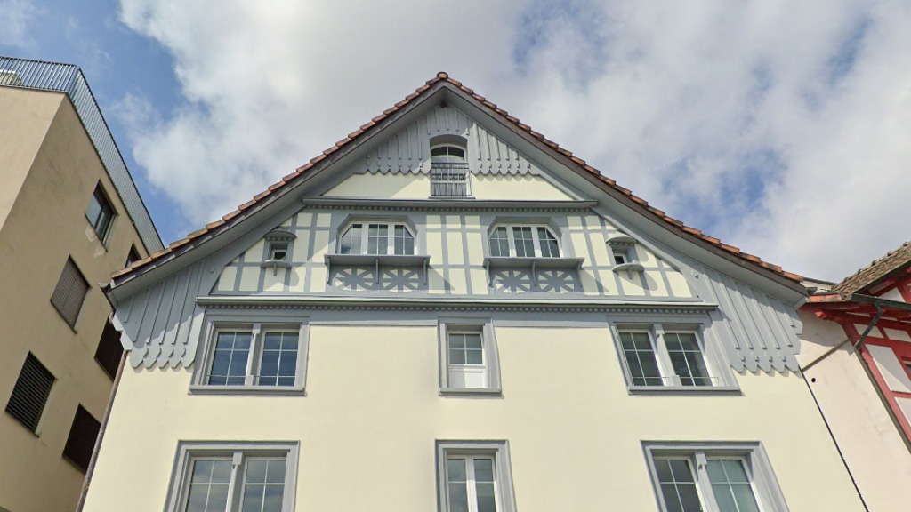 Fasada budynku z charakterystycznym szczytem, kremową elewacją i szarymi elementami dekoracyjnymi, widok z dołu na tle błękitnego nieba z chmurami.