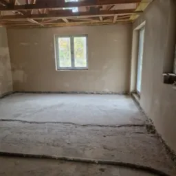 Surowe wnętrze w trakcie prac wykończeniowych: ściany pokryte świeżym tynkiem, widoczne drewniane elementy konstrukcji dachu, okno z widokiem na drzewa, szary betonowy podkład z widoczną rurą...