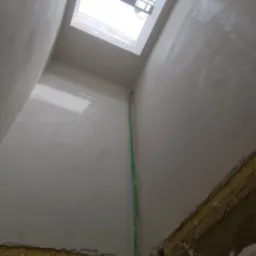 Świeżo wykończone, jasne poddasze z oknem dachowym, widoczne ślady po pracach wykończeniowych na dolnej krawędzi ścian i zwisające kable.