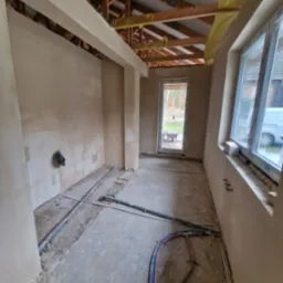 Surowe wnętrze w Lubinie po nałożeniu tynków, widoczne rury na podłodze, okna z PCV i drewniana konstrukcja dachu z izolacją.