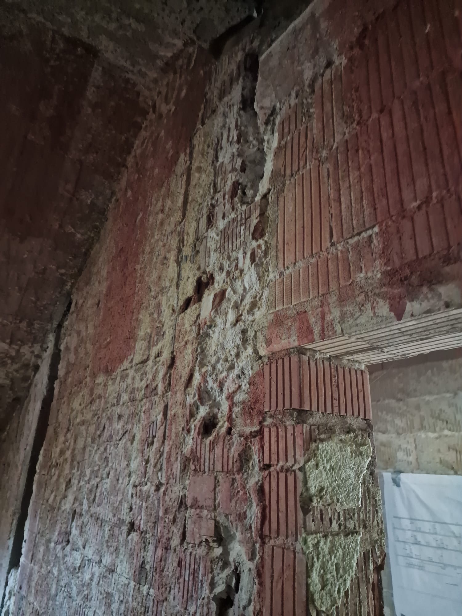 Ceglana ściana w trakcie remontu, z widocznymi śladami starego tynku i fragmentami nowego, jasnego wypełnienia, zarysowany łuk sufitu w tle.