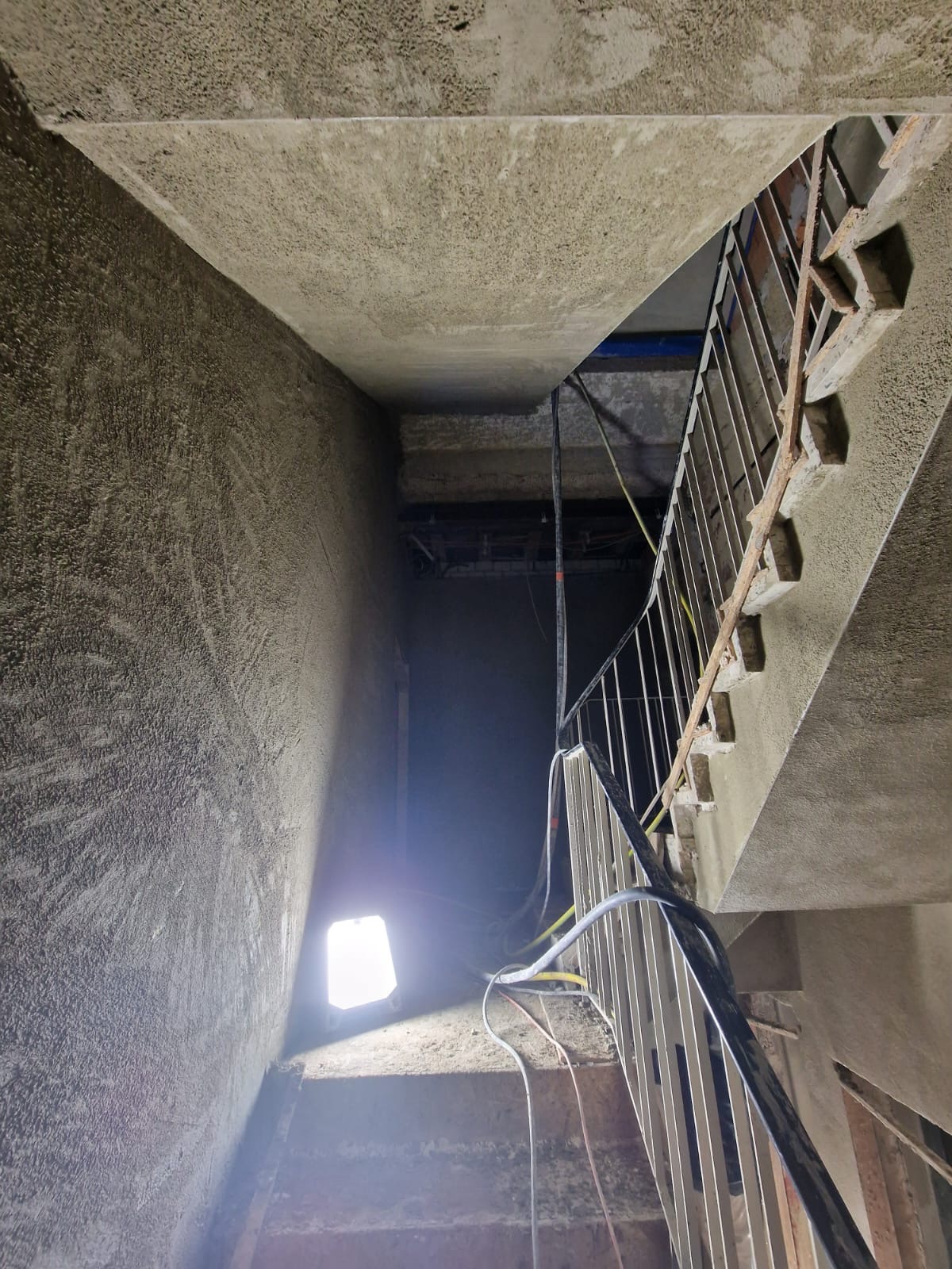 Klatka schodowa w trakcie prac wykończeniowych, ściany pokryte szorstkim tynkiem, widoczne przewody elektryczne i prowizoryczne oświetlenie budowlane.