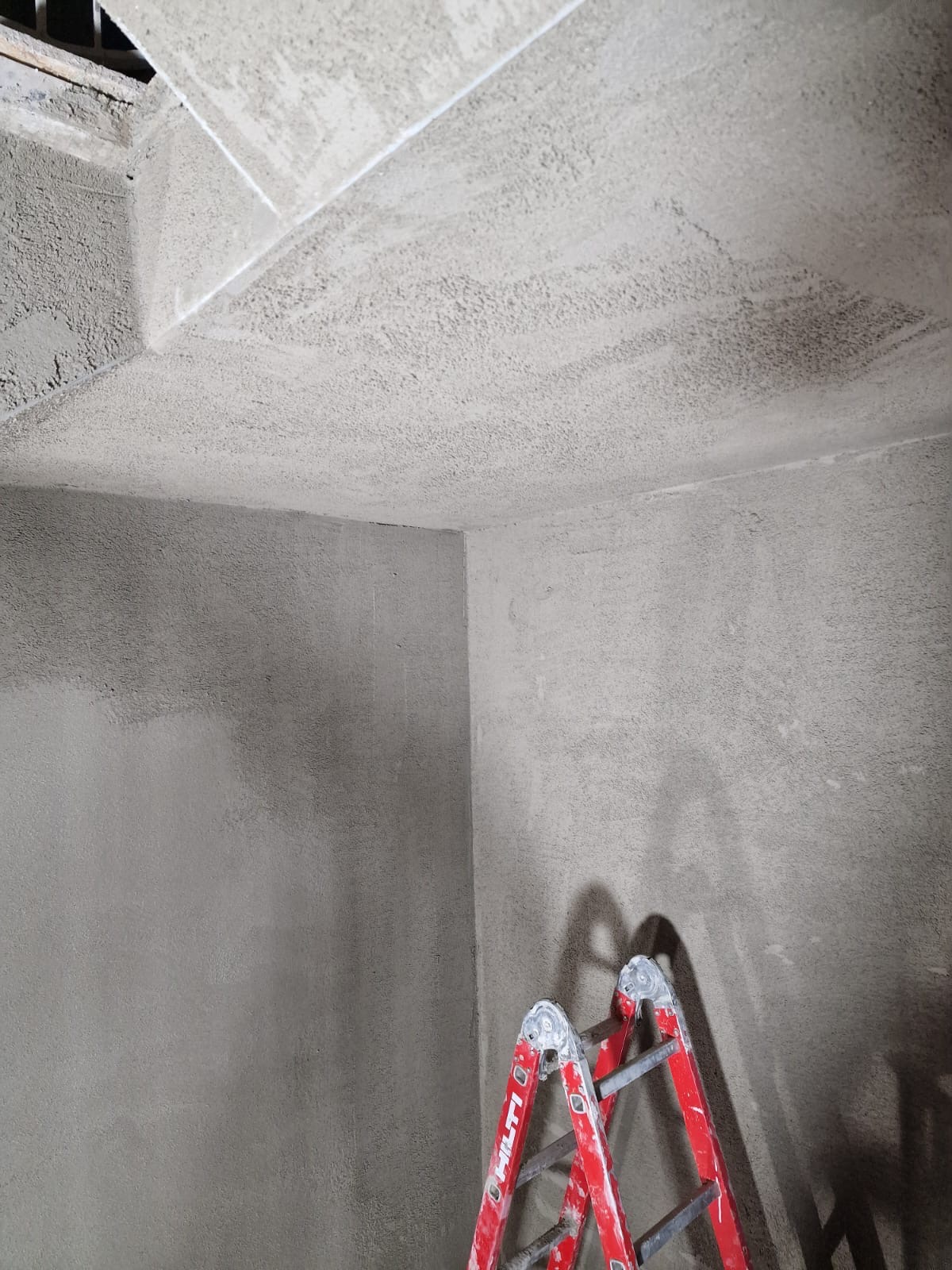 Ściana i sufit po nałożeniu szarej, ziarnistej warstwy, z widoczną czerwoną drabiną Hilti w rogu pomieszczenia.