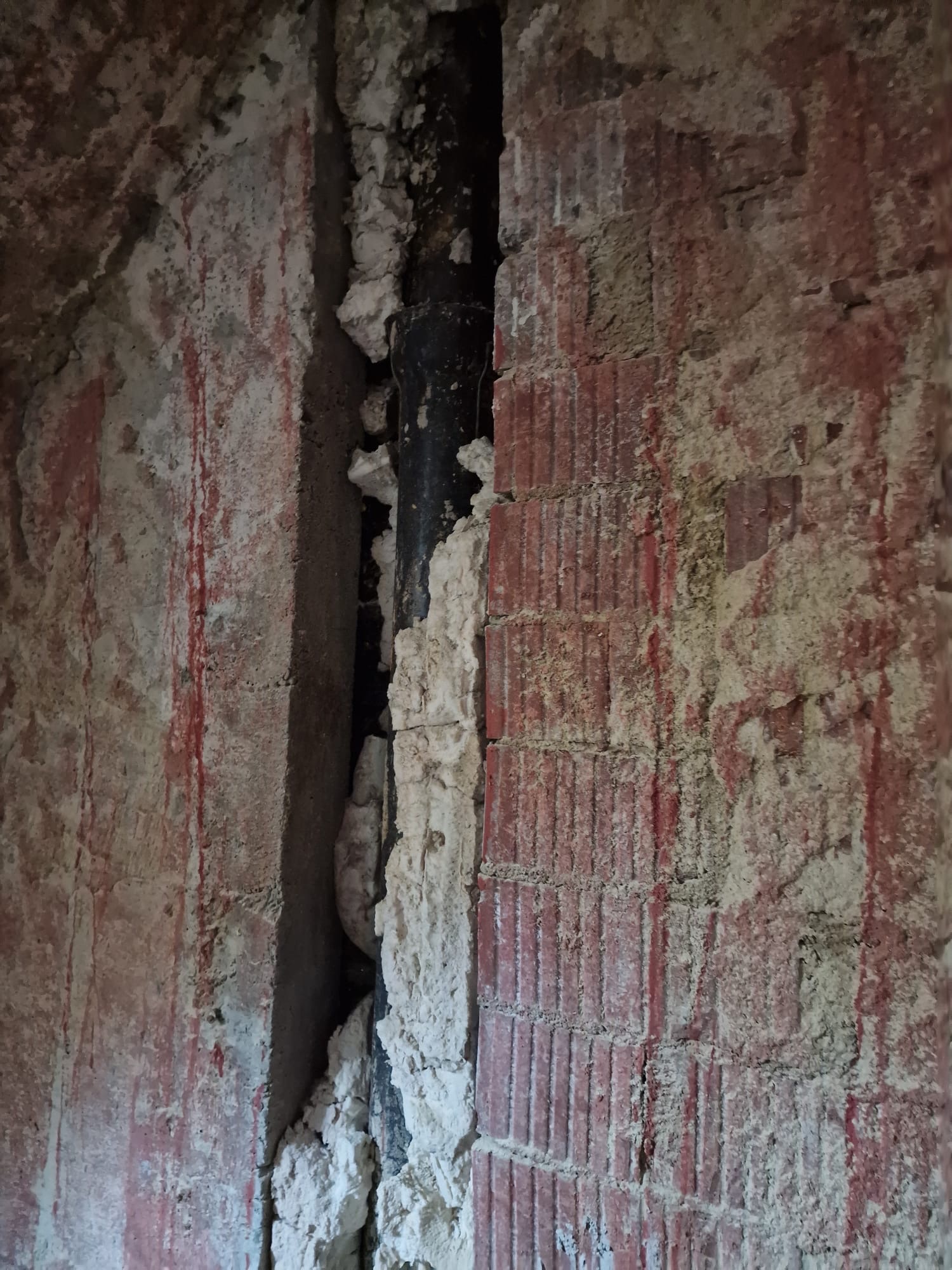 Fragment muru z cegły dziurawki z wyciętym pionowym rowkiem na rurę, wypełnionym częściowo zaprawą gipsową. Widoczne ślady czerwonej farby na ścianie.