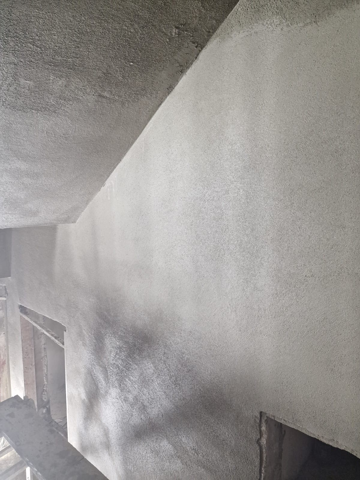 Surowa, szara ściana i sufit pokryte świeżo nałożonym tynkiem, widoczne nierówności faktury i niedoskonałości, z fragmentem ościeżnicy w tle.