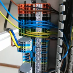 Szczegółowe ujęcie rozdzielnicy elektrycznej z kolorowymi przewodami podłączonymi do listew zaciskowych oznaczonych etykietami, widoczne różne kolory i grubości kabli, a także elementy montażowe...