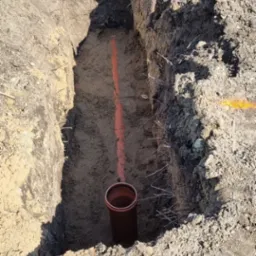 Wykop fundamentowy z widoczną rurą kanalizacyjną i wytyczonym pomarańczową farbą przebiegiem instalacji podziemnej, widoczne ślady pracy koparki.