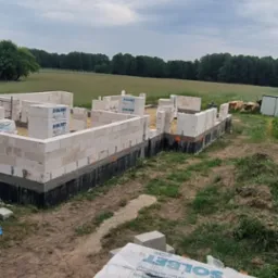 Wznoszenie ścian parteru domu z bloczków Solbet, widoczne fundamenty zabezpieczone izolacją, w tle pole i las pod pochmurnym niebem.