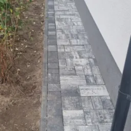 Ułożona ścieżka z kostki brukowej w odcieniach szarości, z widocznym odpływem liniowym przy ścianie budynku o jasnej elewacji i rabatą z roślinami po drugiej stronie.