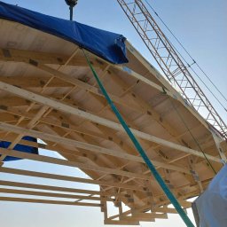 Raf-Bud - Montaż drewnianej konstrukcji dachu domu szkieletowego za pomocą dźwigu, zabezpieczonej niebieską plandeką, widoczni pracownicy w odblaskowych kamizelkach.