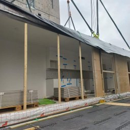 Raf-Bud - Montaż modułowej konstrukcji budynku z gotową kuchnią; widoczne podwieszenie dźwigiem, ściany z płyt warstwowych, drewniane podpory, palety z płytami, izolacja podłogowa z nadrukiem Rothoblaas.