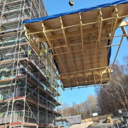 Raf-Bud - Żuraw przenosi prefabrykowany element konstrukcyjny na placu budowy, widoczne rusztowania i budynek w trakcie wznoszenia.