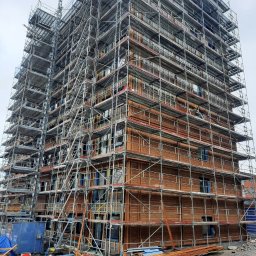 Raf-Bud - Wielopiętrowy budynek w trakcie budowy, obłożony drewnianymi panelami i otoczony rusztowaniami, widoczne materiały budowlane na placu budowy.