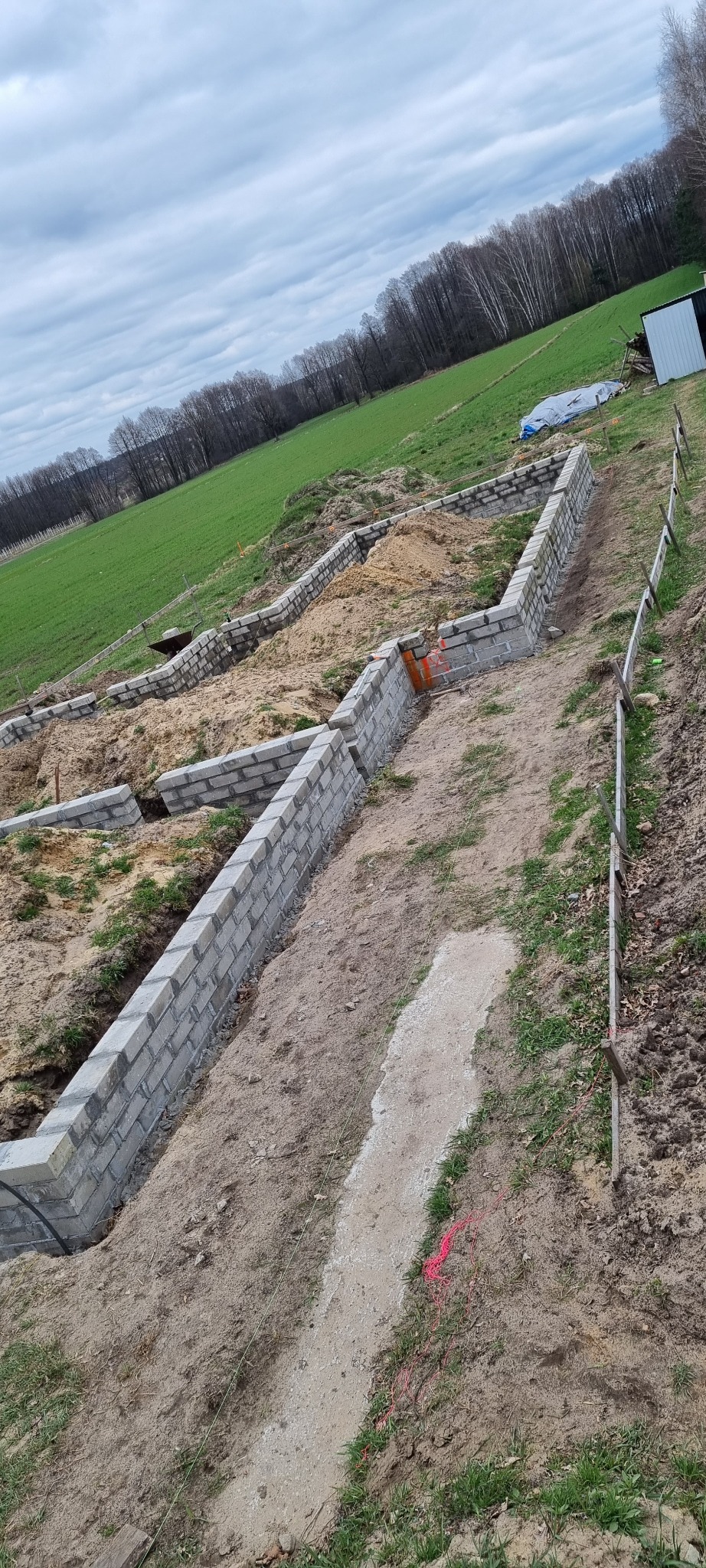 Wykop pod fundamenty z częściowo wymurowanymi ścianami z bloczków betonowych, widoczne wytyczone sznurki i teren budowy z zielonym polem w tle.