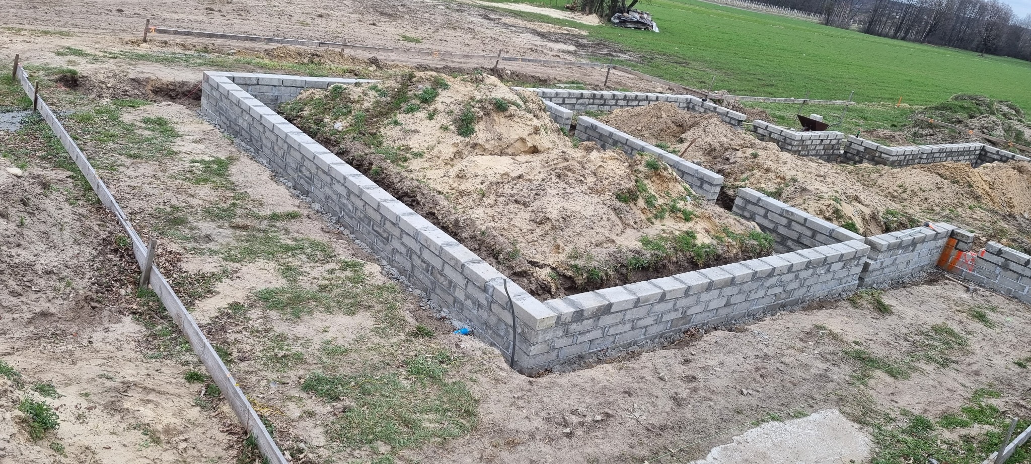 Wznoszone mury fundamentowe z bloczków betonowych, częściowo zasypane ziemią, widoczne drewniane szalunki, teren budowy z zielenią w tle.