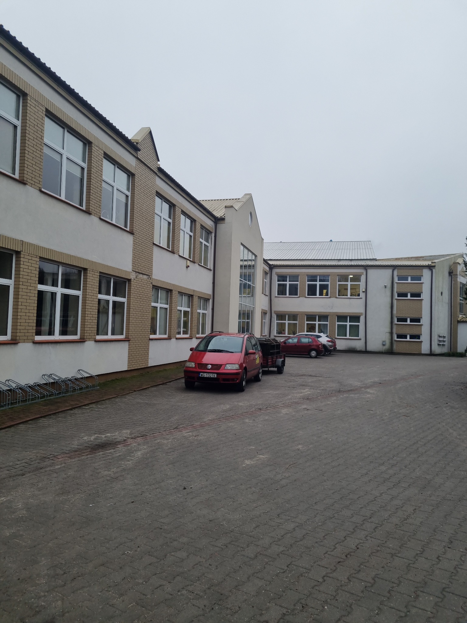 Dziedziniec budynku z kostki brukowej z zaparkowanymi samochodami i stojakami na rowery w pochmurny dzień.