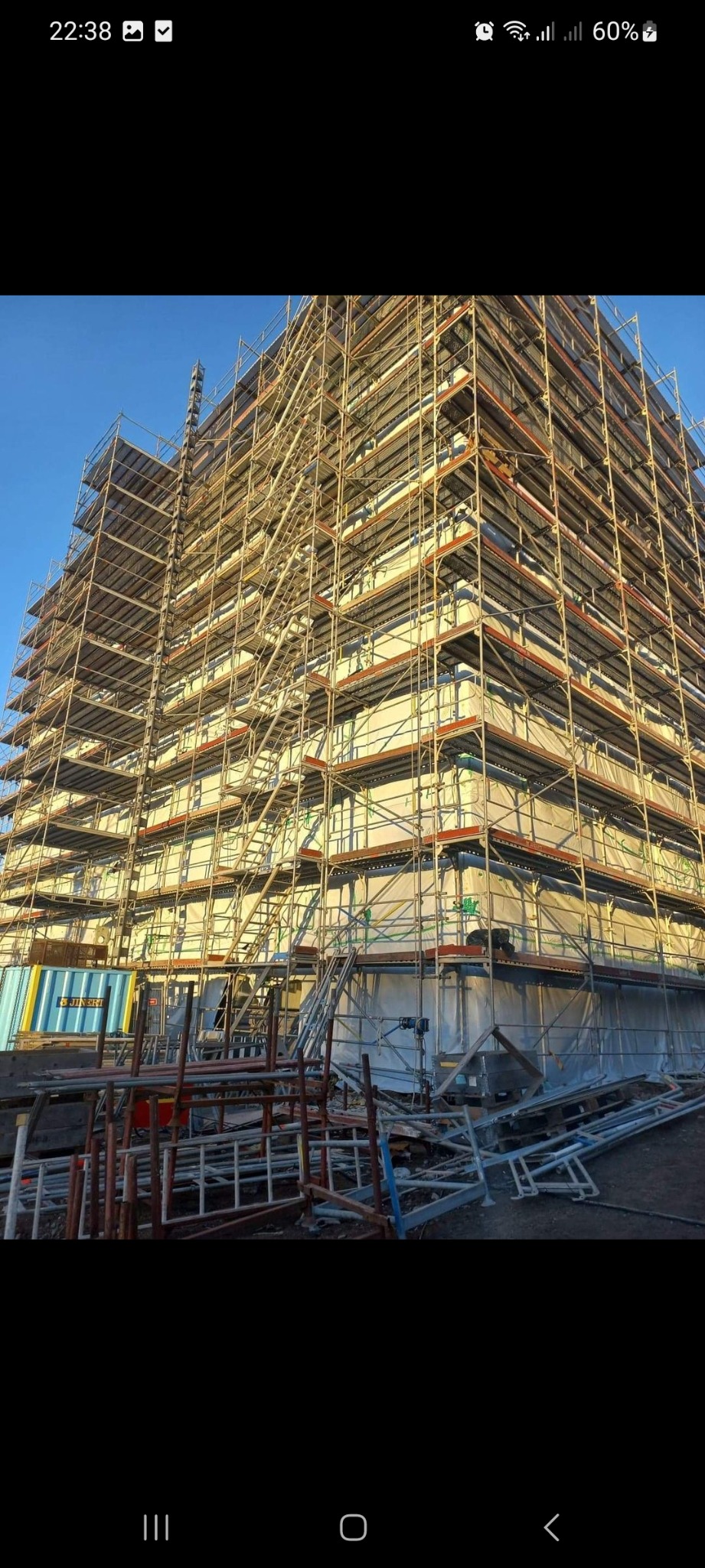 Wysoki budynek w trakcie budowy, całkowicie otoczony rusztowaniami, z widocznym kontenerem i elementami konstrukcyjnymi na pierwszym planie.