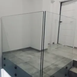 Minimalistyczna szklana balustrada z czarnymi mocowaniami na tle szarej podłogi i białych ścian, widok z g&oacute;ry schod&oacute;w.