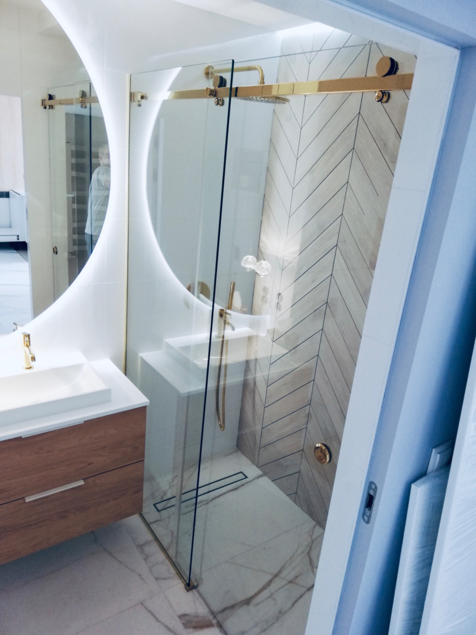 Nowoczesna łazienka z kabiną prysznicową ze szklanymi przesuwnymi drzwiami i złotymi okuciami, widoczna umywalka z drewnianą szafką i okrągłe lustro z podświetleniem LED.