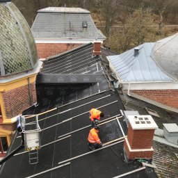 Krystian Gaik - Prace na dachu zabytkowego budynku: dekarze w pomarańczowych kombinezonach montują membrany dachowe Flexilight i drewniane łaty, widoczne kominy i sąsiednie dachy pokryte dachówką, podnośnik...