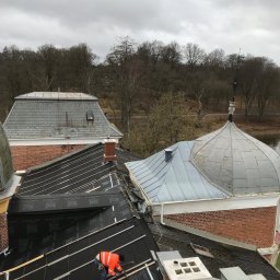 Krystian Gaik - Dwóch dekarzy w pomarańczowych kurtkach montuje czarną membranę dachową na drewnianej konstrukcji dachu, z widokiem na inne dachy z cegły i blachy oraz drzewa w tle.