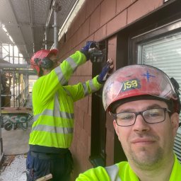Krystian Gaik - Pracownik w hełmie robiący selfie na tle montowanych brązowych płytek elewacyjnych na ścianie budynku, drugi pracownik na rusztowaniu w tle wierci płytki.