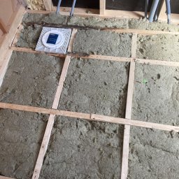 Krystian Gaik - Montaż izolacji z wełny mineralnej między drewnianymi legarami poddasza, widoczne szare rury instalacyjne i biała kratka wentylacyjna