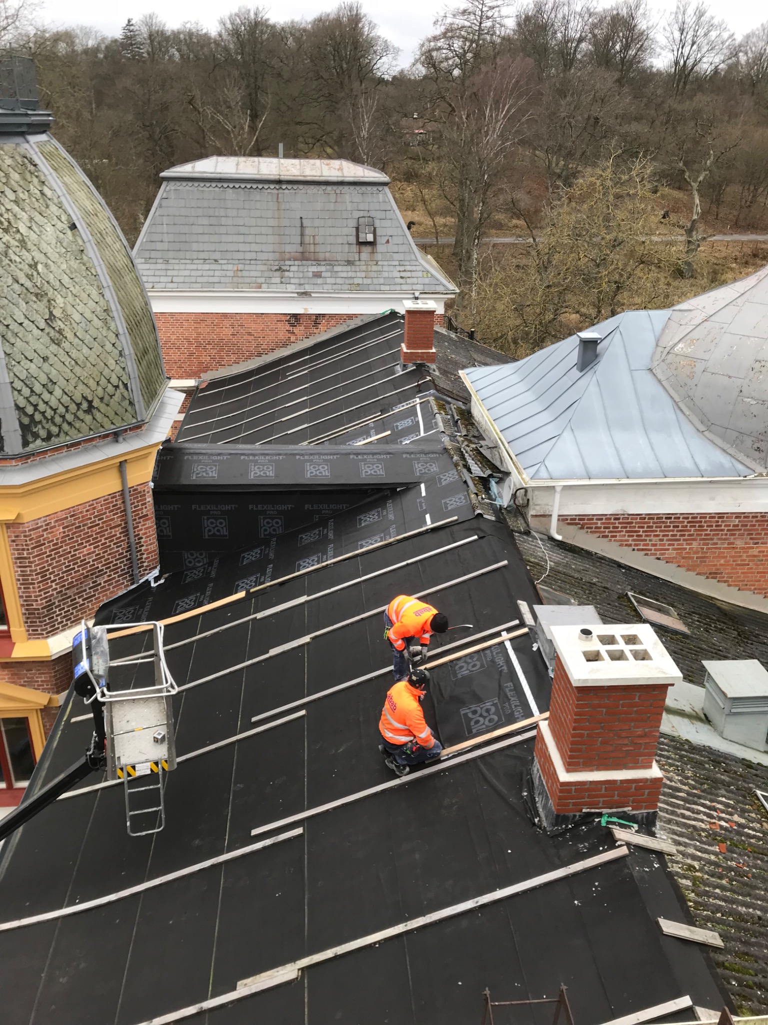Prace na dachu zabytkowego budynku: dekarze w pomarańczowych kombinezonach montują membrany dachowe Flexilight i drewniane łaty, widoczne kominy i sąsiednie dachy pokryte dachówką, podnośnik...
