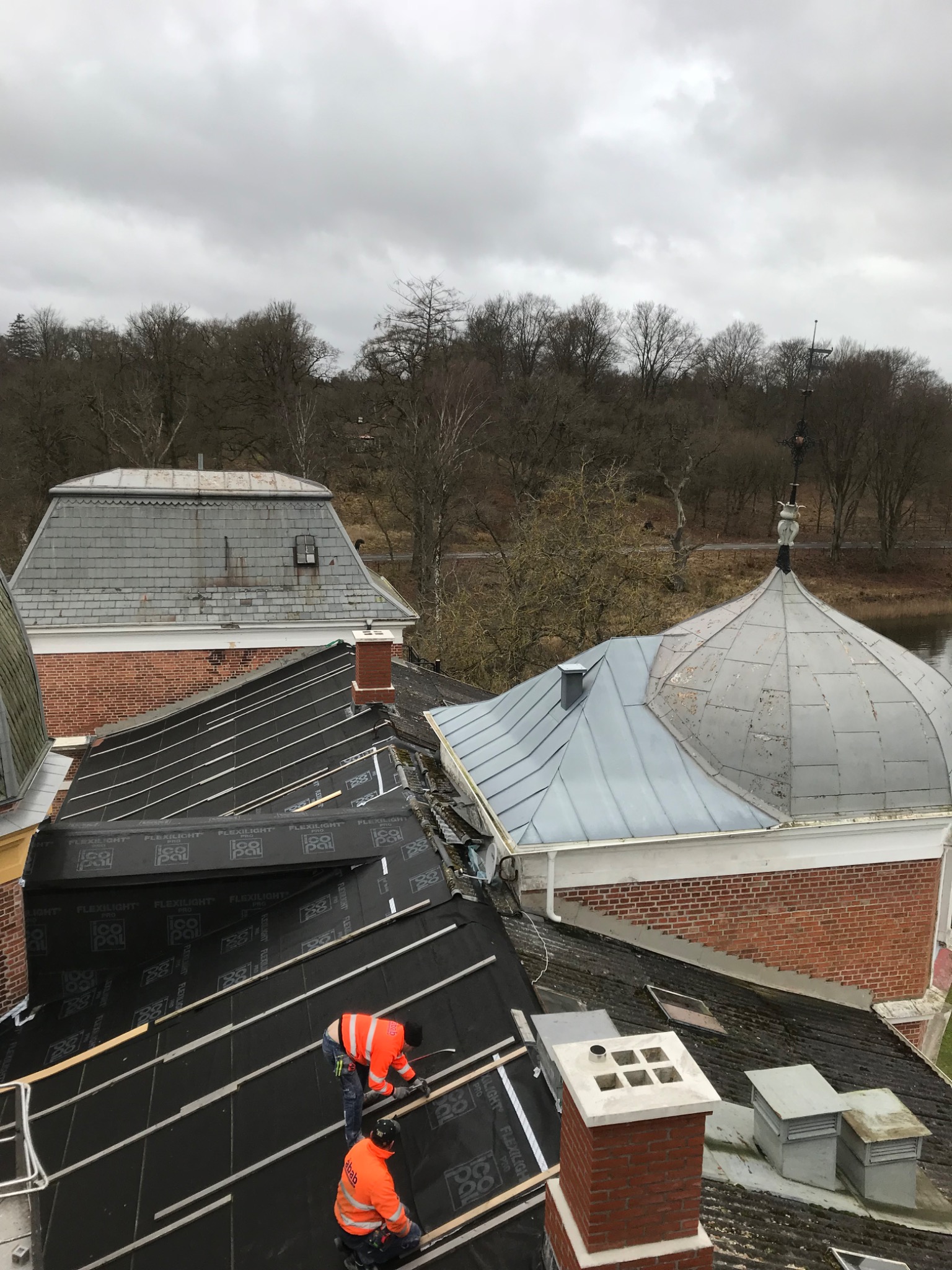 Dwóch dekarzy w pomarańczowych kurtkach montuje czarną membranę dachową na drewnianej konstrukcji dachu, z widokiem na inne dachy z cegły i blachy oraz drzewa w tle.