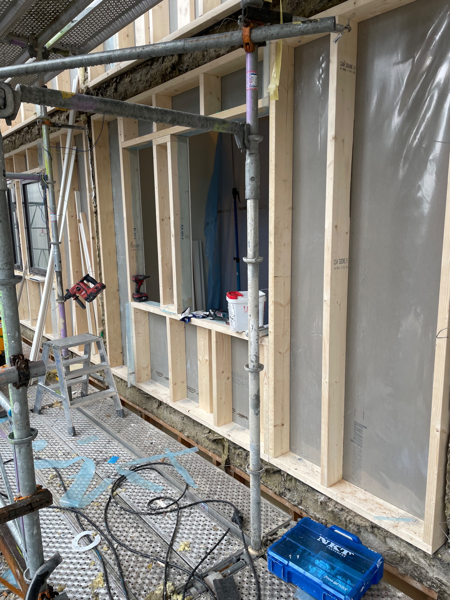 Budowa szkieletu domu z drewnianych belek, widoczne rusztowanie, narzędzia, okna oraz niebieska skrzynka z narzędziami NKT Fasteners