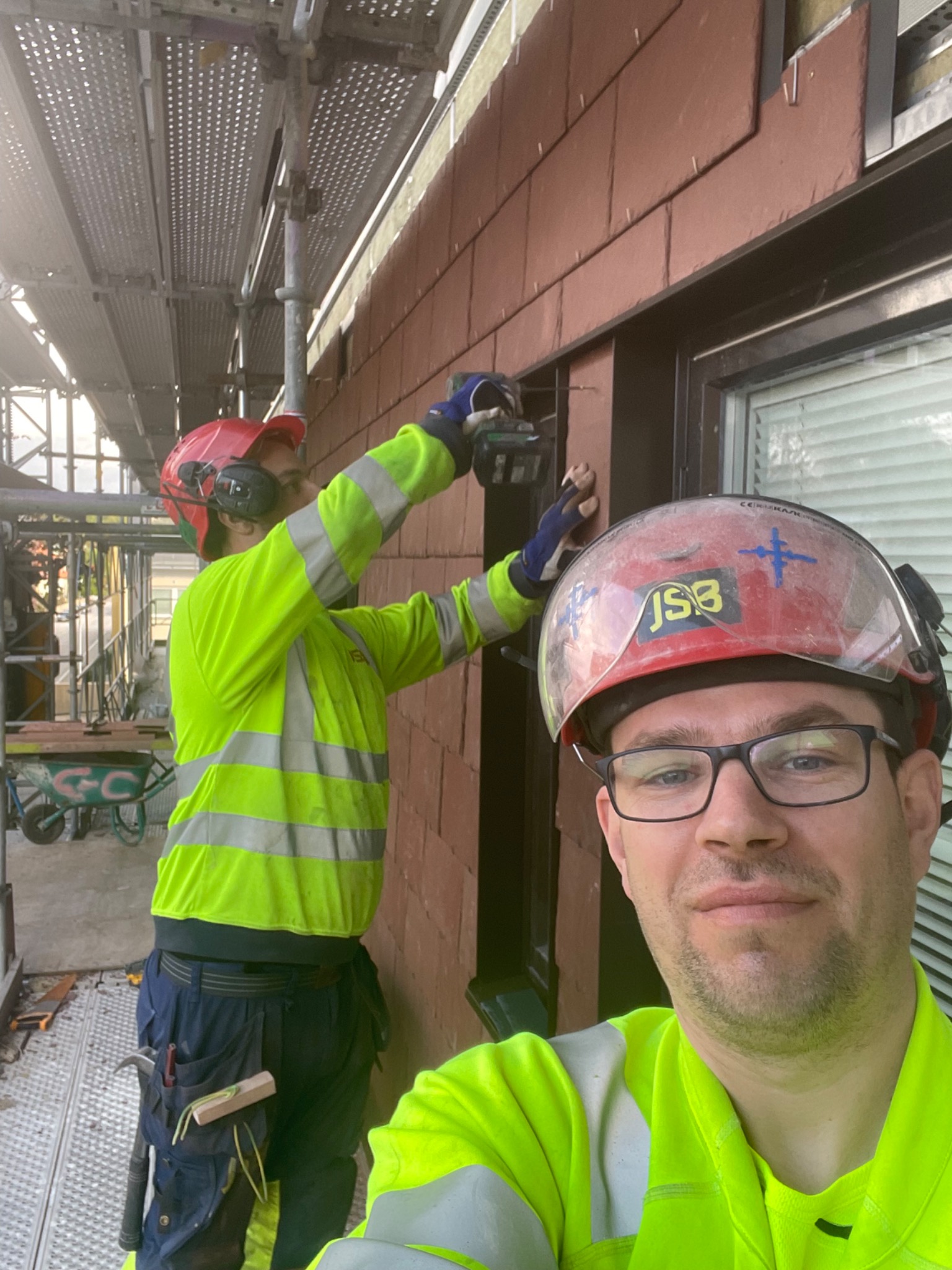 Pracownik w hełmie robiący selfie na tle montowanych brązowych płytek elewacyjnych na ścianie budynku, drugi pracownik na rusztowaniu w tle wierci płytki.